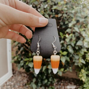 May include: A pair of dangle earrings featuring a candy corn design. The earrings are made of white, orange, and yellow polymer clay and have silver hooks.