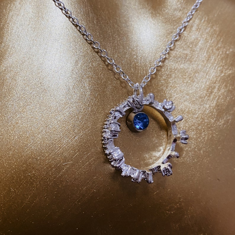 Op de afbeelding: Een zilveren ketting met een ronde hanger met een blauwe edelsteen en een rij kleine heldere stenen rond de rand.