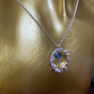Op de afbeelding: Een zilveren ketting met een kroonhanger met een blauwe edelsteen. De kroon is gemaakt van kleine, heldere stenen.