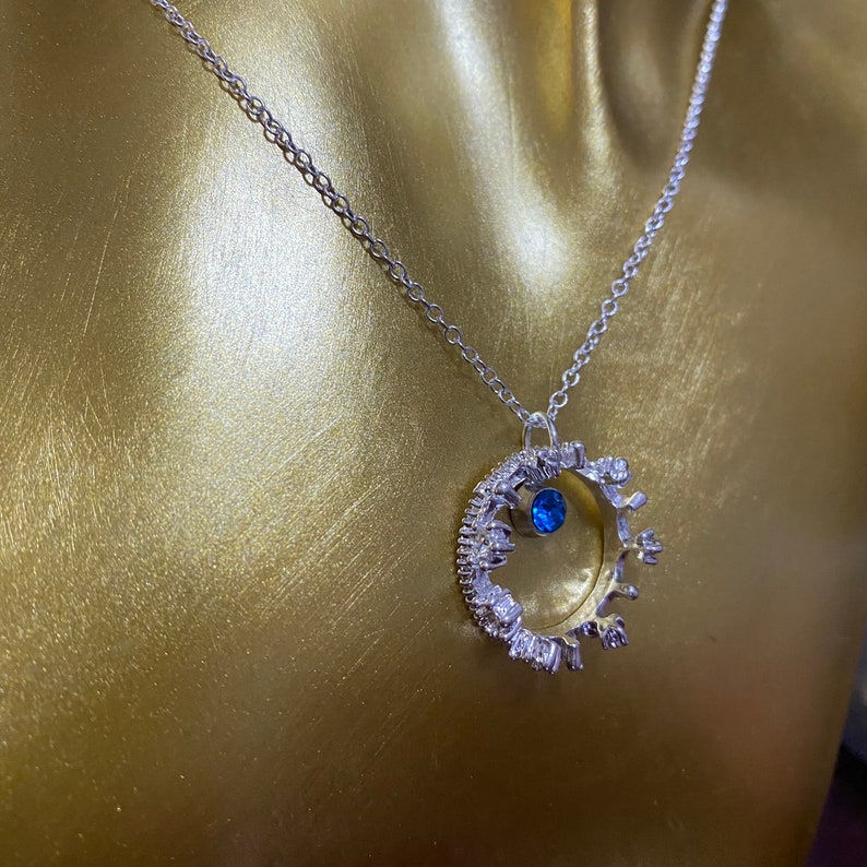Op de afbeelding: Een zilveren ketting met een kroonhanger met een blauwe edelsteen. De kroon is gemaakt van kleine, heldere stenen.