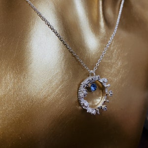 Op de afbeelding: Een zilveren ketting met een ronde hanger met een blauwe edelsteen omringd door kleinere stenen. De hanger is in de vorm van een kroon.