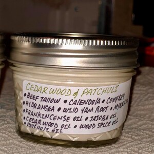 Può includere: Un piccolo barattolo di vetro con coperchio argentato, riempito con una sostanza cremosa. Un'etichetta bianca avvolge il barattolo, elencando ingredienti come legno di cedro, patchouli e vari oli. Il testo è in verde e nero.