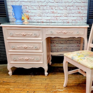 May include: A light pink vanity desk with a glass top, three drawers, and a matching chair. The desk features crystal-style knobs and floral carvings on the drawer fronts. The chair has a spindle back and a seat upholstered in floral fabric.