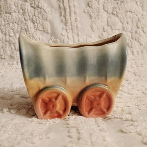 May include: A vintage ceramic planter shaped like a wagon with blue, yellow, and orange stripes. The wagon has two wheels with a detailed design.