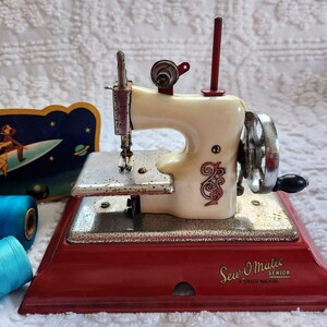 May include: A vintage Sew-O-Matic Senior sewing machine in cream and red. The machine has a silver hand wheel and needle, with decorative scrollwork. A retro sign with a rocket ship and spools of blue thread are in the background.