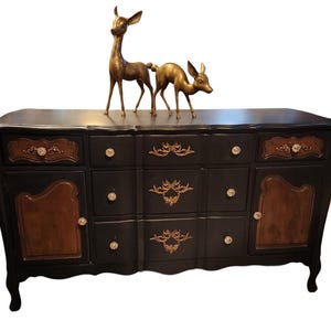 May include: A black antique dresser with gold accents and a brown wood panel design. The dresser has three drawers and two cabinets with crystal knobs. Two gold deer figurines are on top.
