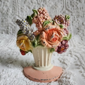 May include: A cast iron doorstop featuring a bouquet of painted flowers in shades of pink, yellow, purple, and white. The flowers are arranged in a fluted, cream-colored vase set upon a semi-circular, light pink base with a distressed finish.