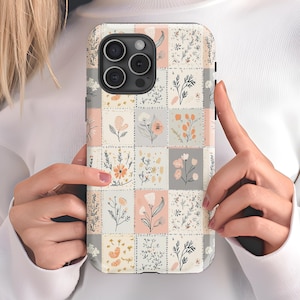 May include: A phone case with a patchwork design featuring a variety of floral patterns in shades of pink, white, and gray. The case has a black border and a cutout for the phone's camera.