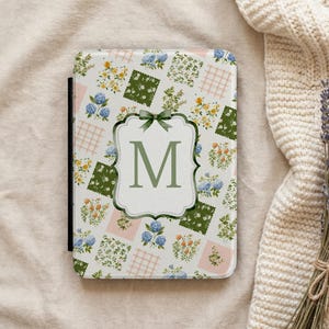 May include: A floral patterned tablet case with the letter "M" in a green frame. The case features a patchwork design with blue, yellow, and green floral squares, and pink and white grid squares. A green bow adorns the top of the frame.