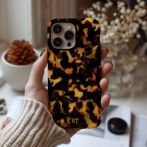 May include: A smartphone case featuring a dark brown, black, and amber tortoiseshell pattern. The case has a cutout for a triple-lens camera system with gold-toned rings and the gold-colored initials BT printed near the bottom edge of the case.
