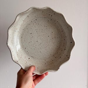 May include: A speckled, off-white ceramic plate with a wavy, scalloped edge. The plate has a shallow depth and is covered in small, dark brown speckles. The plate is held by a hand against a plain white background.