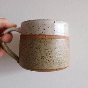 May include: A ceramic mug featuring a two-tone design with a speckled off-white upper section and a speckled light brown lower section. The mug has a curved handle on the left side and a smooth, rounded rim, showcasing a distinct horizontal line.