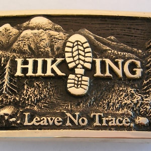 May include: A brass belt buckle with a mountain scene and a boot print. The buckle says "HIKING" and "Leave No Trace".