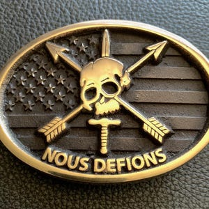 May include: A gold-tone belt buckle with a skull and crossbones design. The buckle features a US flag with stars and stripes, and the words "NOUS DEFIONS" in a banner below the skull.