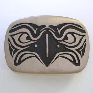 May include: A rectangular, brushed metal belt buckle with a stylized black eagle design. The eagle's face is centered, with detailed wing and eye patterns. The buckle has rounded edges and a matte finish.