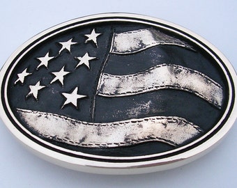 May include: An oval-shaped belt buckle featuring a silver American flag design. The flag is raised and has a black background with white stars and stripes.