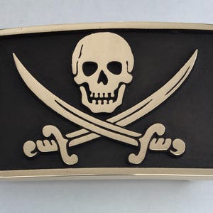 May include: A silver belt buckle with a black background and a white skull and crossbones design.