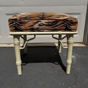 May include: A small, cream-colored bamboo-style stool with a rectangular seat. The seat is upholstered with a brown and black tiger stripe pattern. The stool has four legs and is set against a neutral background.