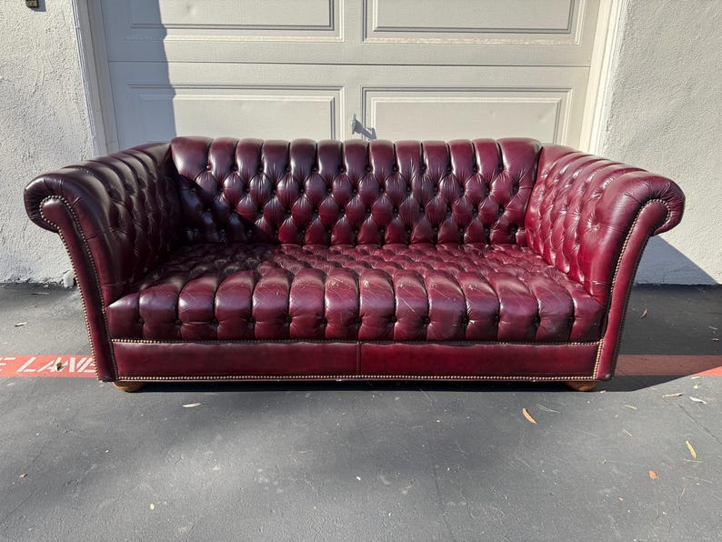 May include: A burgundy leather Chesterfield sofa with a button-tufted design. The sofa has rolled arms and a low profile. The sofa is approximately 84 inches long.