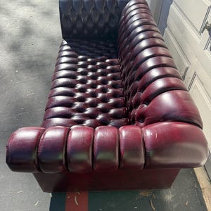 May include: A burgundy leather sofa with a button-tufted back and rolled arms. The sofa has a low profile and a classic design, perfect for a living room or lounge area. The leather has a slight sheen, and the sofa appears to be in good condition.