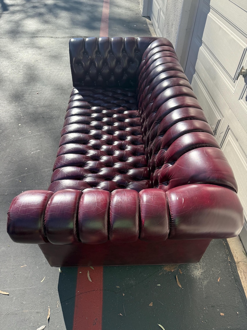 May include: A burgundy leather sofa with a button-tufted back and rolled arms. The sofa has a low profile and a classic design, perfect for a living room or lounge area. The leather has a slight sheen, and the sofa appears to be in good condition.