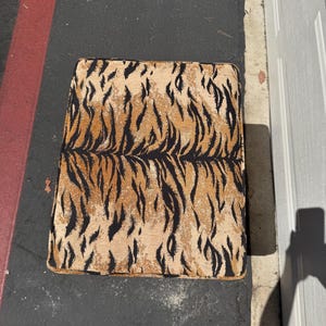 May include: A square ottoman with a tiger stripe pattern in shades of tan, brown, and black. The ottoman is placed on a dark surface, with a red stripe and a white door visible in the background.