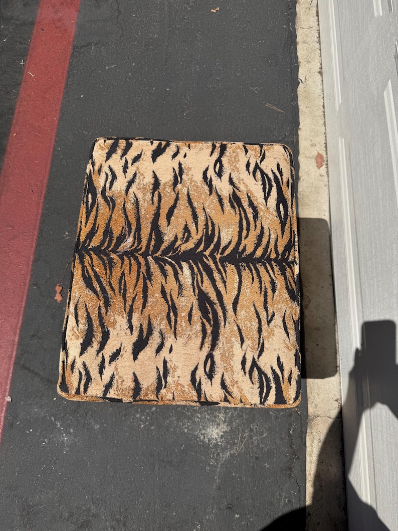 May include: A square ottoman with a tiger stripe pattern in shades of tan, brown, and black. The ottoman is placed on a dark surface, with a red stripe and a white door visible in the background.