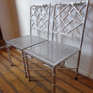 May include: Two silver chairs with a bamboo-style frame and a diamond lattice backrest. The seats are made of perforated metal. The chairs are on a wooden floor. The chairs are a decorative furniture item.
