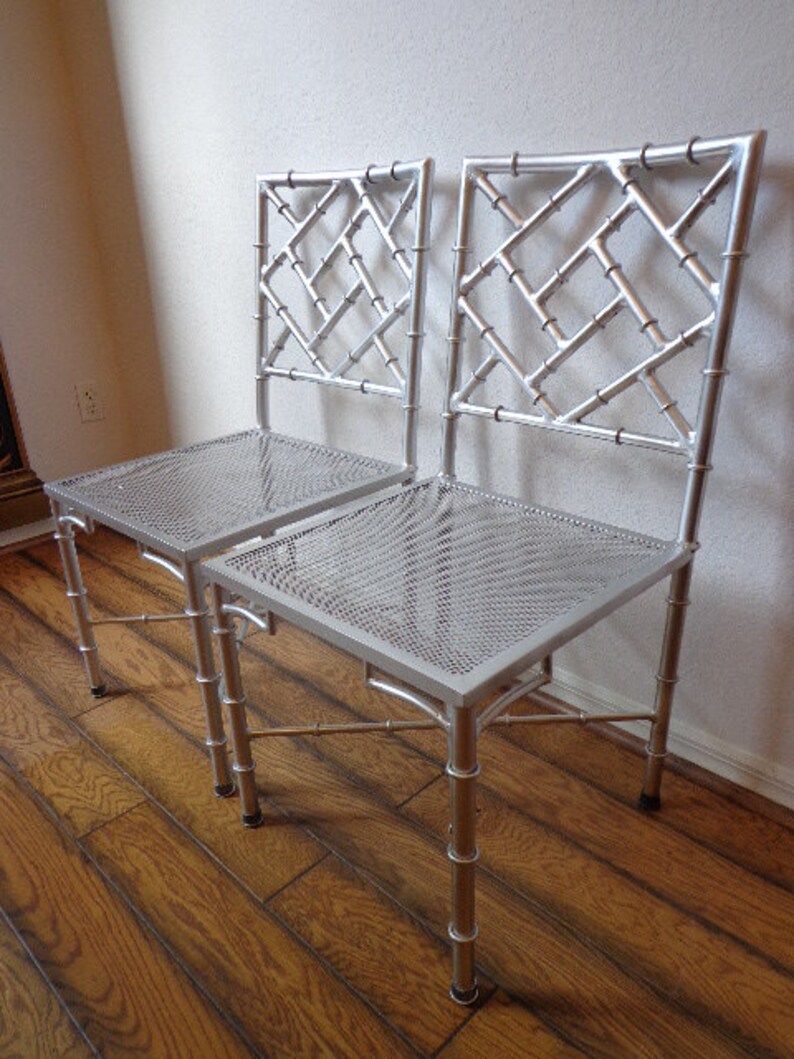 May include: Two silver chairs with a bamboo-style frame and a diamond lattice backrest. The seats are made of perforated metal. The chairs are on a wooden floor. The chairs are a decorative furniture item.