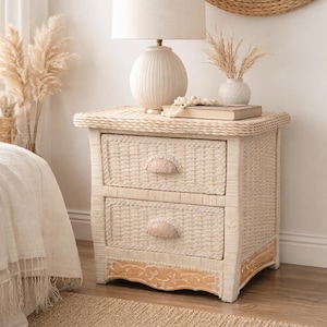 May include: A two-drawer nightstand made of woven wicker in a light beige color. The nightstand has a decorative wooden base and shell-shaped drawer pulls. A lamp with a cream-colored shade sits on top, along with a vase and a book. The room has a neutral color palette.