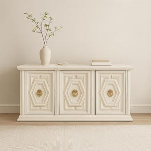 May include: Cream-colored cabinet with three doors, each featuring a geometric design and gold-toned hardware. A white vase with greenery and a small dish sit atop the cabinet. The cabinet is on a cream-colored rug.