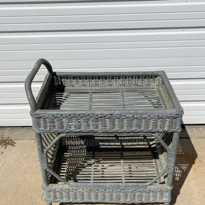 May include: A gray wicker serving cart with two shelves and wheels. The cart has a handle on the top.