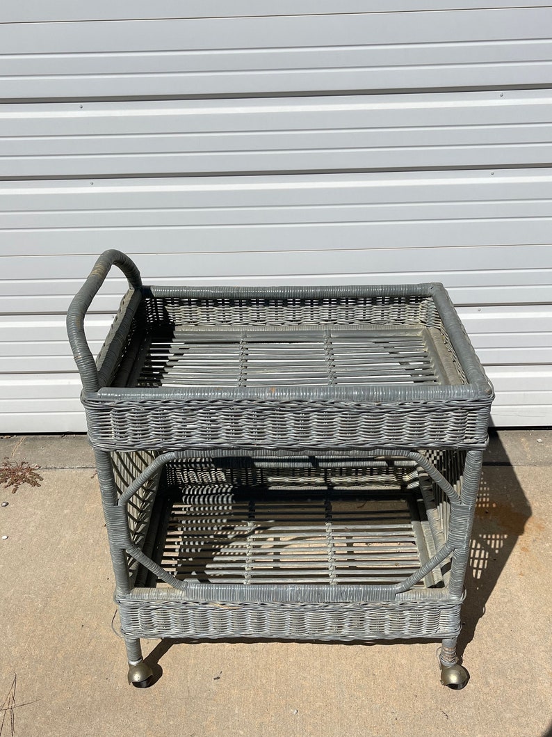 May include: A gray wicker serving cart with two shelves and wheels. The cart has a handle on the top.