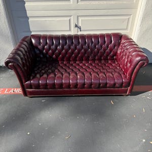 May include: A burgundy leather sofa with a button-tufted design. The sofa has rolled arms and a low profile. The sofa is approximately 84 inches long.