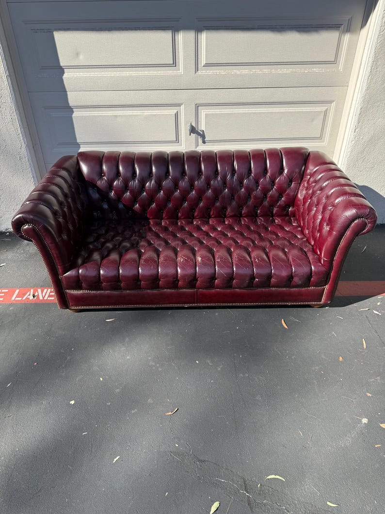 May include: A burgundy leather sofa with a button-tufted design. The sofa has rolled arms and a low profile. The sofa is approximately 84 inches long.