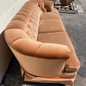 May include: A vintage, tufted, peach-colored velvet sofa with a rounded back and a skirt. The sofa is in good condition and has a classic design.