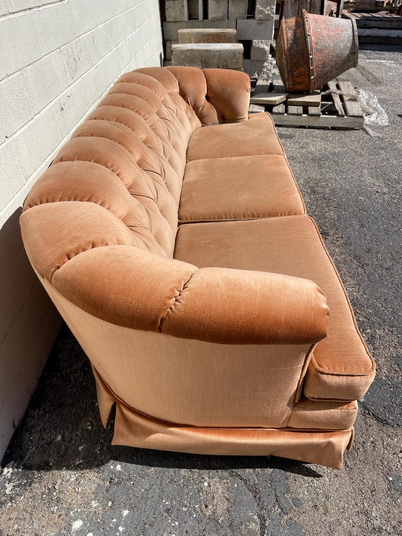 May include: A vintage, tufted, peach-colored velvet sofa with a rounded back and a skirt. The sofa is in good condition and has a classic design.