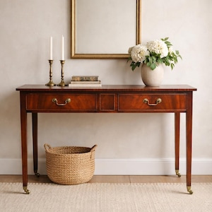 May include: A mahogany console table with two drawers, brass hardware, and caster wheels. A gold-framed mirror hangs above the table, with candles, books, and a vase of white flowers. A woven basket sits below on a neutral rug.