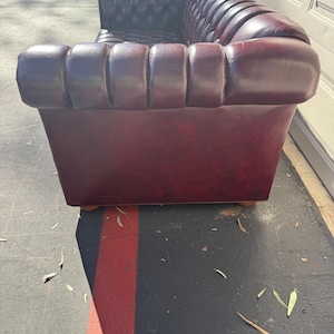 May include: A burgundy leather Chesterfield sofa with a tufted back and rolled arms. The sofa has a classic design with a rectangular shape and wooden feet. The leather appears to be in good condition, with a rich color and subtle sheen.