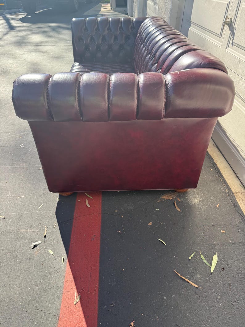 May include: A burgundy leather Chesterfield sofa with a tufted back and rolled arms. The sofa has a classic design with a rectangular shape and wooden feet. The leather appears to be in good condition, with a rich color and subtle sheen.