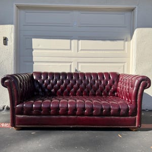 May include: A burgundy leather Chesterfield sofa with button tufting. The sofa has rolled arms and sits on wooden feet. The background features a white garage door.