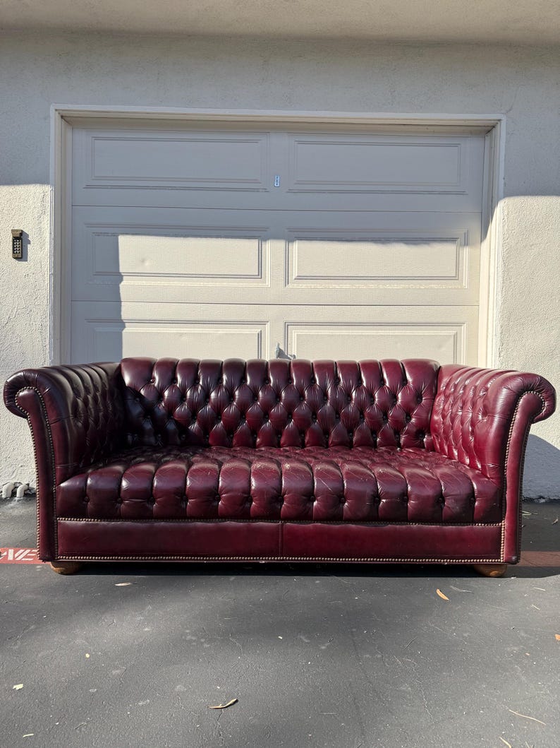 May include: A burgundy leather Chesterfield sofa with button tufting. The sofa has rolled arms and sits on wooden feet. The background features a white garage door.