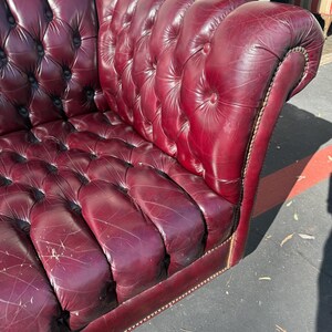 May include: A burgundy leather Chesterfield sofa with button tufting. The sofa has a curved back and armrests, and is trimmed with a decorative nailhead border. The leather shows signs of wear, adding to its vintage appeal.