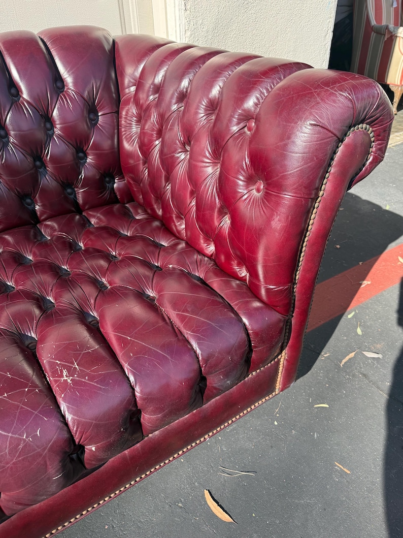 May include: A burgundy leather Chesterfield sofa with button tufting. The sofa has a curved back and armrests, and is trimmed with a decorative nailhead border. The leather shows signs of wear, adding to its vintage appeal.