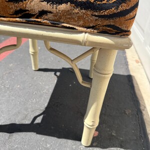 May include: A cream-colored bamboo-style bench with a tiger-striped cushion. The bench has four legs and a curved support structure. The cushion features a brown and black pattern.
