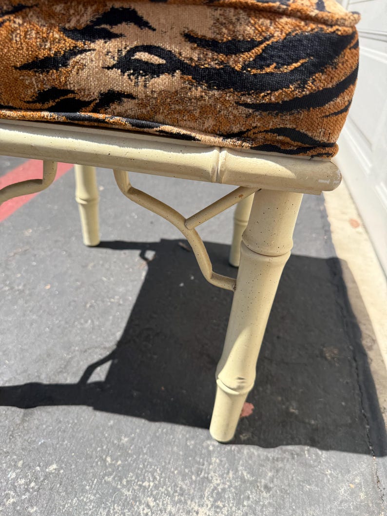 May include: A cream-colored bamboo-style bench with a tiger-striped cushion. The bench has four legs and a curved support structure. The cushion features a brown and black pattern.