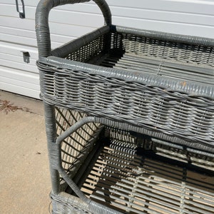 May include: A gray wicker serving cart with two shelves and a handle. The cart is made of woven wicker and has a distressed finish.
