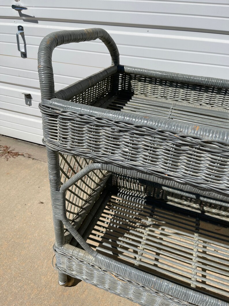 May include: A gray wicker serving cart with two shelves and a handle. The cart is made of woven wicker and has a distressed finish.