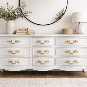 May include: A white dresser with nine drawers and ornate gold-colored handles. The dresser has a curved apron and cabriole legs. A vase with greenery, a mirror, and a lamp are on top.