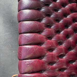 May include: Close-up of a burgundy leather upholstered surface with a tufted design. The leather shows signs of wear and tear, with visible scratches and indentations. The surface is likely part of a piece of furniture, such as a sofa or headboard.